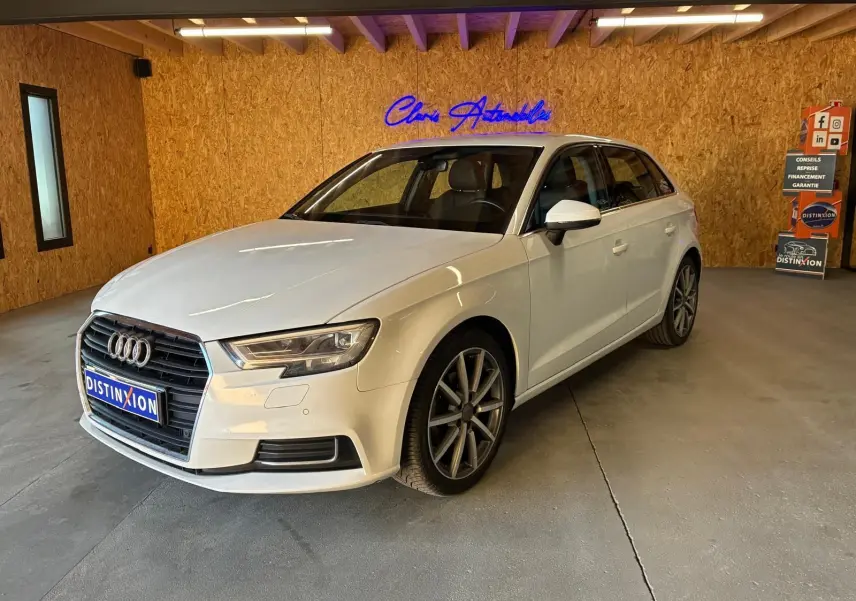 Audi A3 Sportback blanc vue 3/4 avant droit dans un intérieur avec mur en bois et enseigne lumineuse bleue.