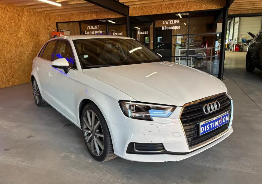 Audi A3 Sportback blanc vue 3/4 avant droit dans un atelier avec calandre noire et jantes alliage.