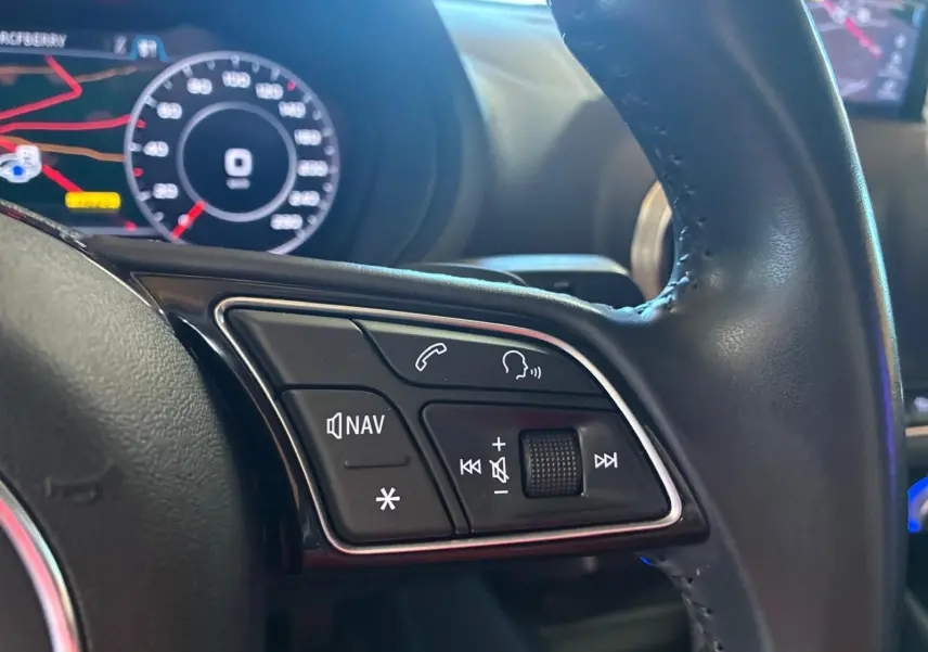 Gros plan sur les commandes audio et navigation au volant en cuir noir de l'Audi A3 Sportback blanc, tableau de bord numérique en arrière-plan.