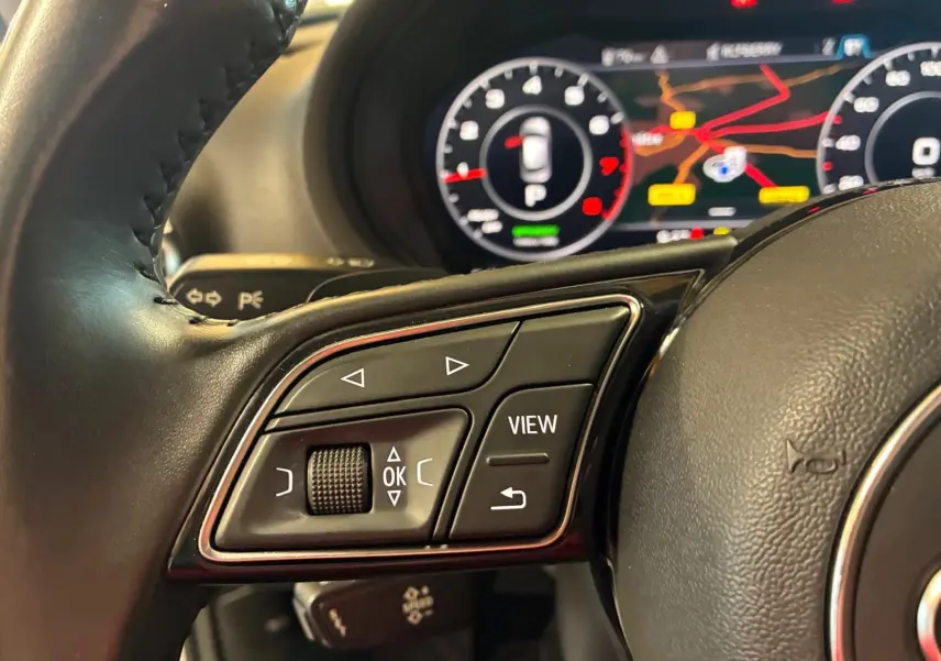 Gros plan sur les commandes au volant côté gauche de l'Audi A3 Sportback blanc avec tableau de bord numérique en arrière-plan.