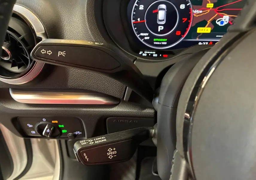 Gros plan sur le volant et le combiné d'instruments numérique d'une Audi A3 Sportback blanche, affichant la navigation et les commandes au volant.