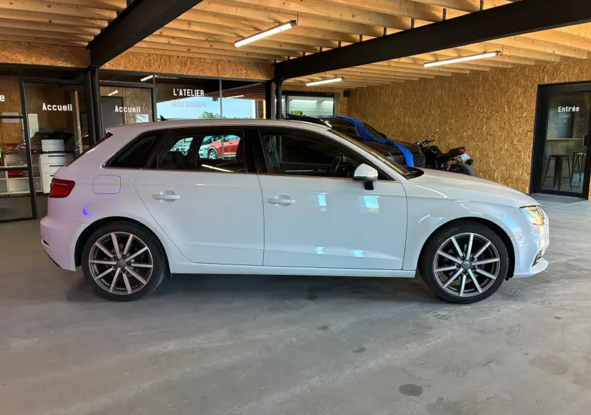 Audi A3 Sportback blanc vue de profil côté gauche, stationnée dans un garage avec intérieur en bois.