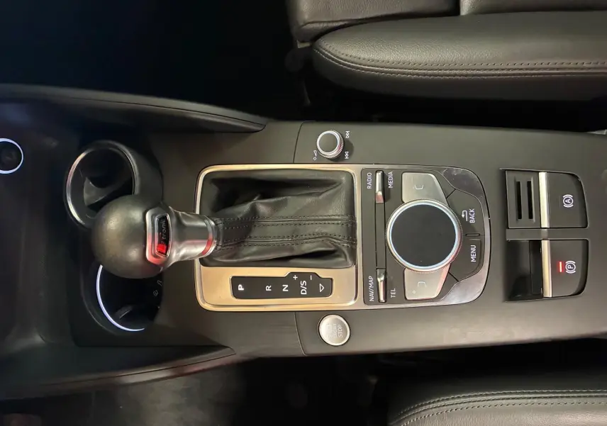 Vue rapprochée du levier de vitesses automatique et du système de commande multimédia sur la console centrale noire d’une Audi A3 Sportback blanche.