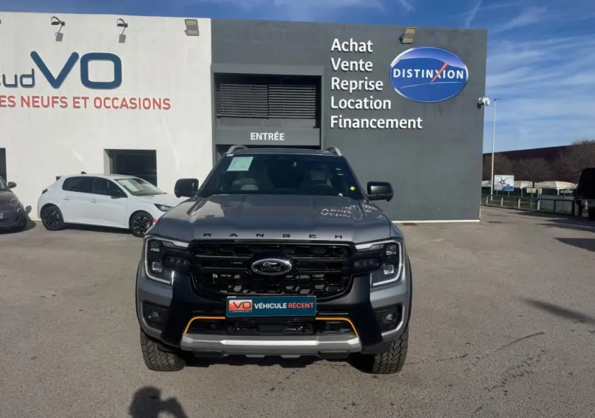 Vue de face d'un Ford Ranger gris 2025 avec calandre noire et barres de toit, stationné devant un concessionnaire.