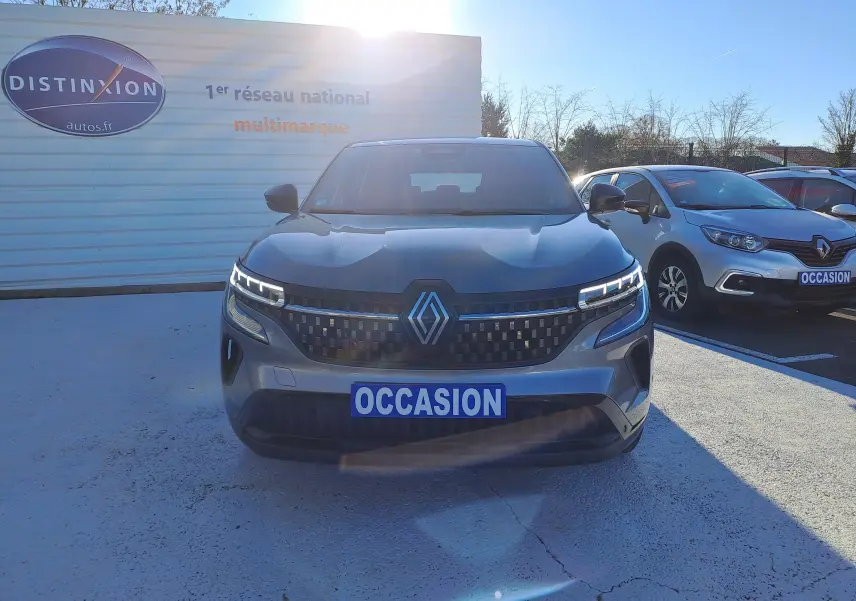 Vue de face d'un Renault Austral gris schiste 2023 avec calandre à damiers et phares LED allumés, sur parking extérieur.