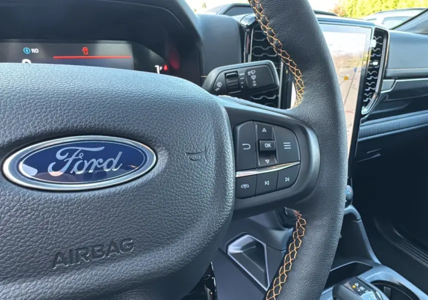 Gros plan sur le volant en cuir noir avec surpiqûres orange du Ford Ranger PHEV 281 Wildtrak, tableau de bord et écran tactile visibles.