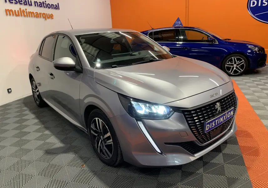 Peugeot 208 gris vue 3/4 avant droit avec phares allumés, calandre noire et jantes alliage en intérieur showroom.