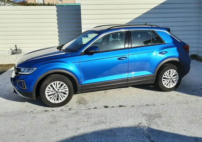 Volkswagen T-Roc bleu en profil côté gauche, avec jantes alliage et toit noir sur fond urbain lumineux.