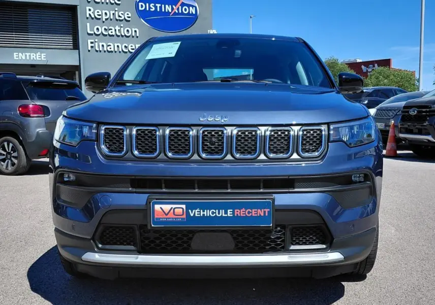 Vue frontale d'un Jeep Compass bleu 2025 avec calandre chromée et plaque "Véhicule récent" sur parking.