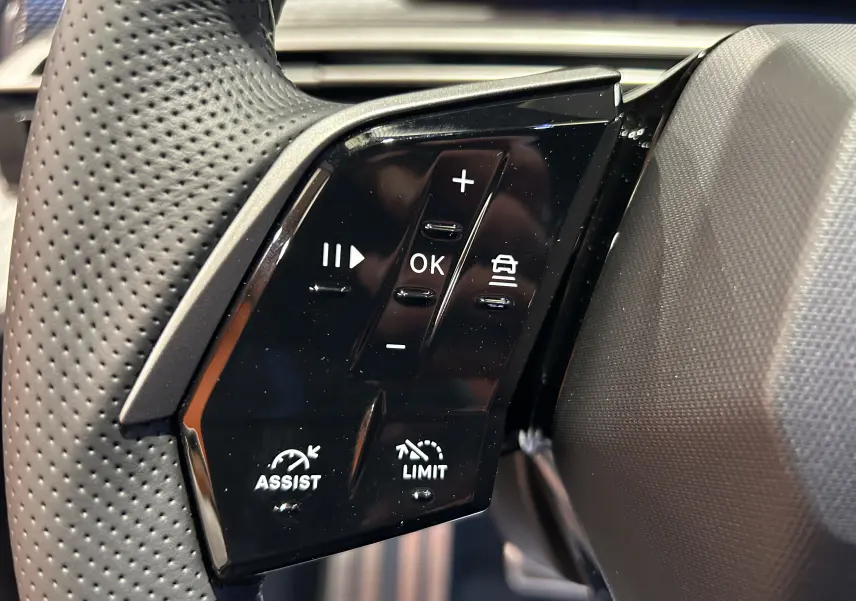 Gros plan sur les commandes au volant noir perforé du Peugeot 5008 III GT Hybrid 145 e-DCS6, avec boutons d'assistance et limitation.