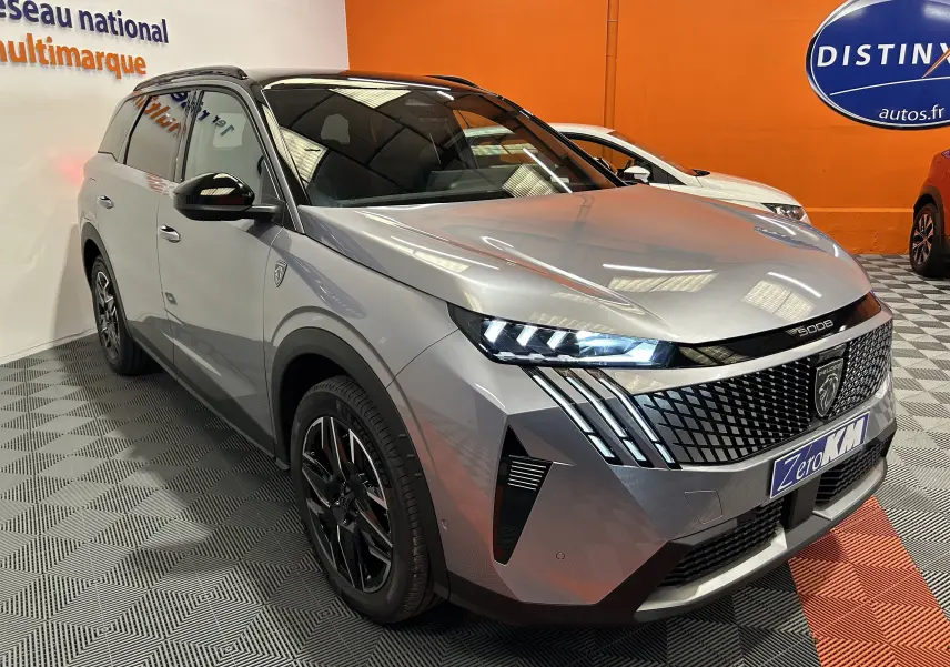 Peugeot 5008 III GT Hybrid gris Artense en 3/4 avant droit, avec calandre distinctive et phares LED allumés.