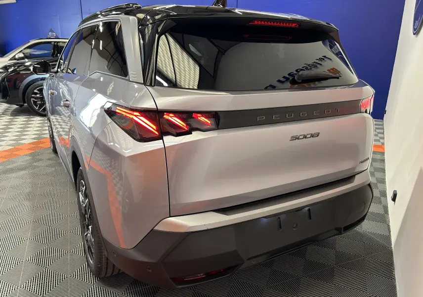 Vue 3/4 arrière droite du Peugeot 5008 gris Artense avec feux LED allumés et toit noir brillant en intérieur showroom.