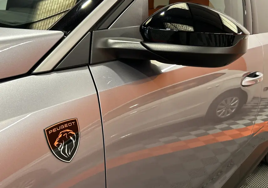 Gros plan sur le côté gauche gris Artense du Peugeot 5008 III GT Hybrid 145 avec logo lion noir et orange sur l'aile.