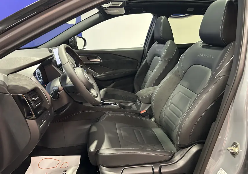 Vue intérieure côté conducteur du Nissan Qashqai gris céramique, sièges cuir noir avec logo Qashqai brodé et toit panoramique ouvert.