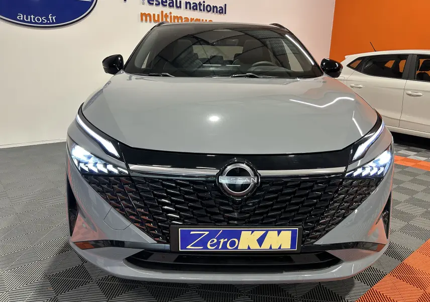 Vue de face du Nissan Qashqai gris céramique métal avec calandre noire et phares LED allumés dans un showroom.