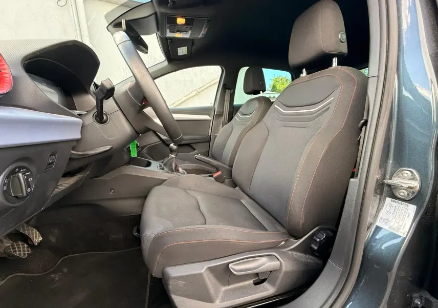 Vue intérieure côté conducteur de la SEAT Ibiza 1.0 TSI 95 FR 2025 en gris foncé, avec sièges tissu noirs et coutures rouges.