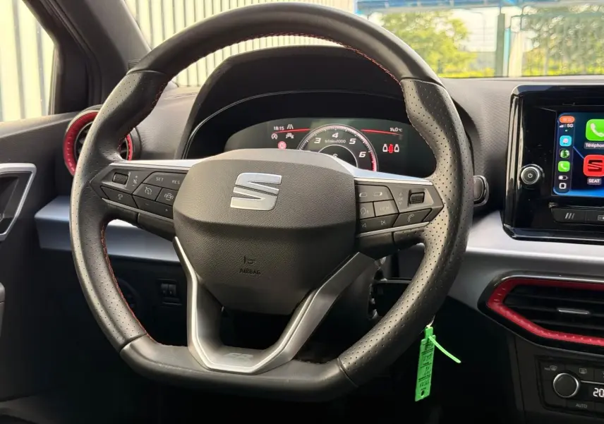 Vue intérieure centrée sur le volant perforé noir avec logo SEAT, tableau de bord numérique et écran tactile à droite.
