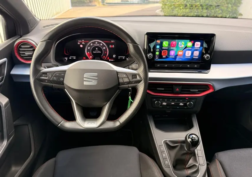 Vue intérieure frontale du poste de conduite de la SEAT Ibiza 1.0 TSI 95 FR 2025, volant sport, écran tactile et levier de vitesses manuel.