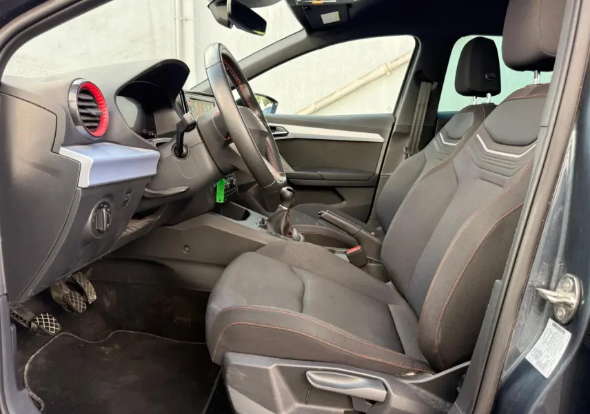 Vue intérieure côté conducteur de la SEAT Ibiza 1.0 TSI 95 FR 2025 avec sièges gris foncé et surpiqûres rouges.