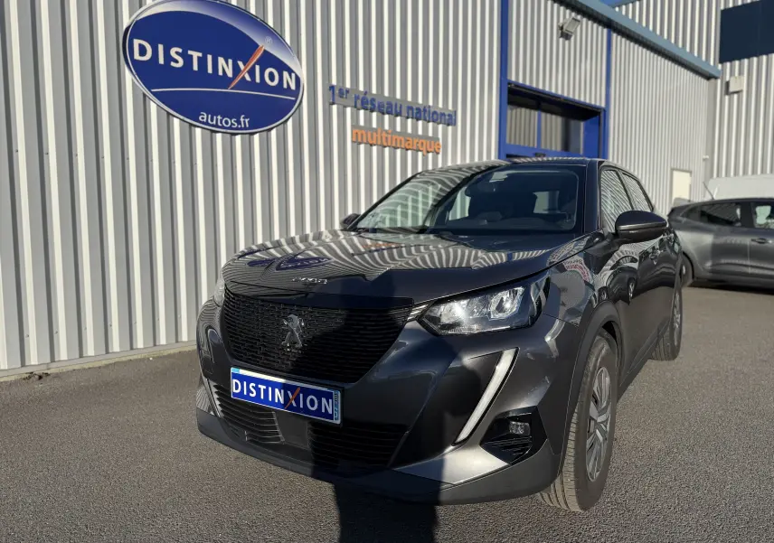 Peugeot 2008 gris 3/4 avant droit, avec calandre noire et phares LED, stationnée devant un bâtiment industriel.
