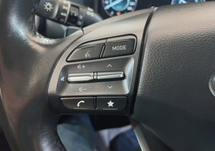 Gros plan sur les commandes gauche du volant en cuir noir du Hyundai Kona 1.6 Hybrid 2022, boutons multifonctions visibles.