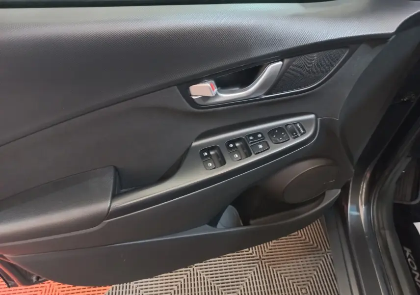 Vue intérieure côté gauche de la porte avant gris foncé du Hyundai Kona 1.6 Hybrid avec commandes électriques et poignée métal.