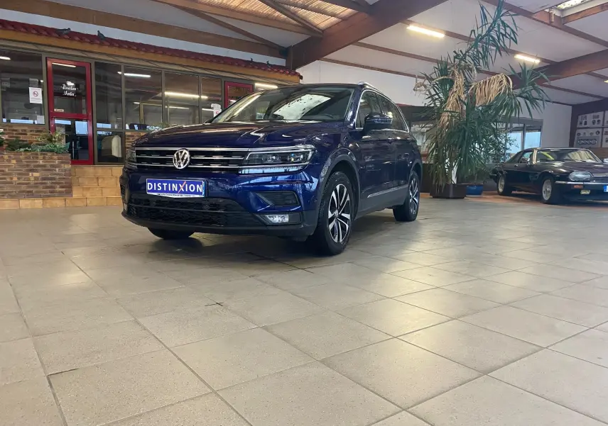 Volkswagen Tiguan bleu vert en 3/4 avant droit dans un showroom avec sol carrelé et plantes en arrière-plan.