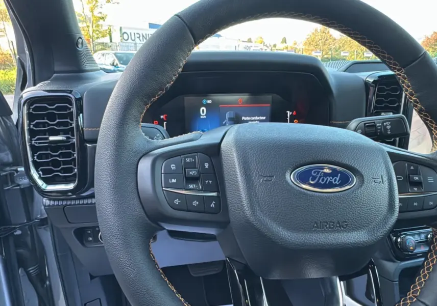 Vue rapprochée du volant cuir noir avec surpiqûres orange du Ford Ranger PHEV 281 Wildtrak 2025, tableau de bord numérique visible.