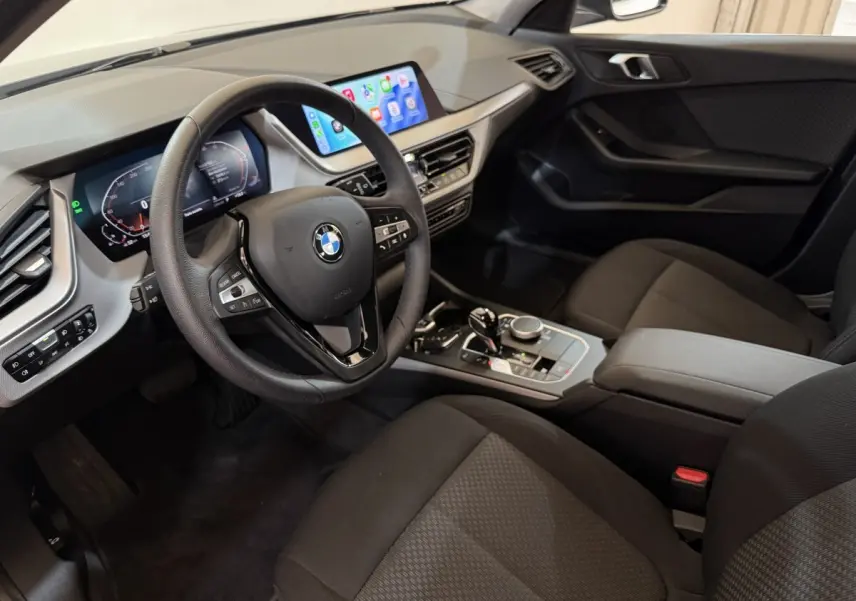 Vue intérieure côté conducteur de la BMW Série 1 116i 2023, tableau de bord moderne avec écran tactile et volant cuir noir.