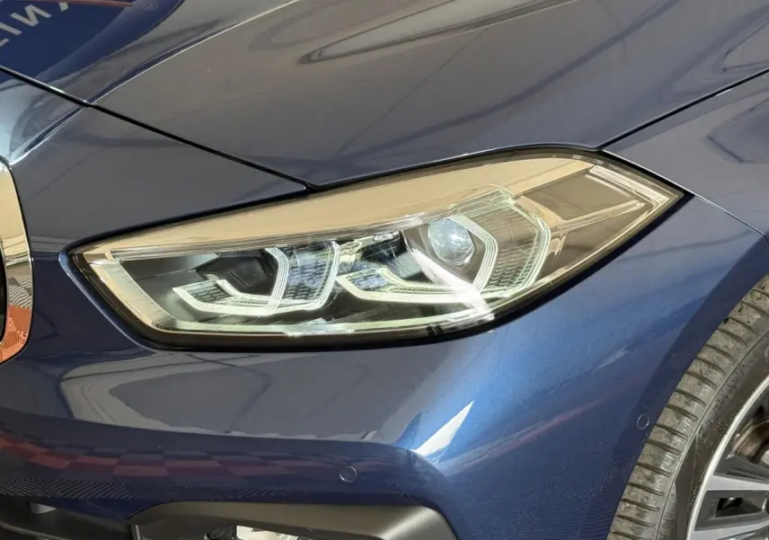 Gros plan sur le phare avant droit d'une BMW Série 1 116i bleu métallisé avec détails LED et partie de la roue visible.