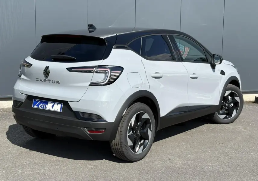 Vue 3/4 arrière droit d'un Renault Captur blanc avec toit noir et jantes noires sur fond gris urbain.