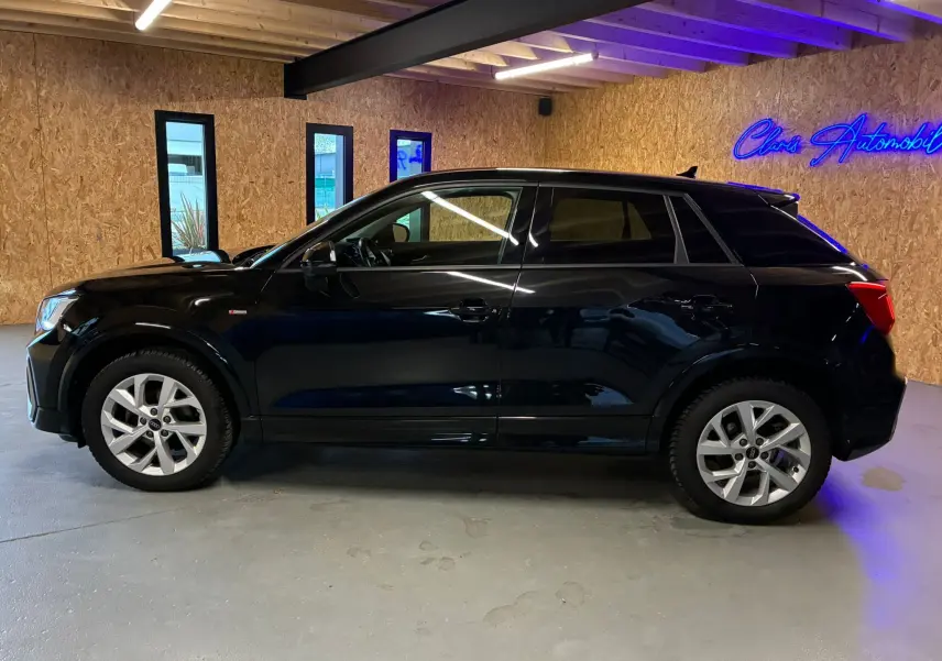 Profil côté gauche d'un Audi Q2 noir 2023 S-Line, avec jantes alliage et vitres teintées dans un garage éclairé.