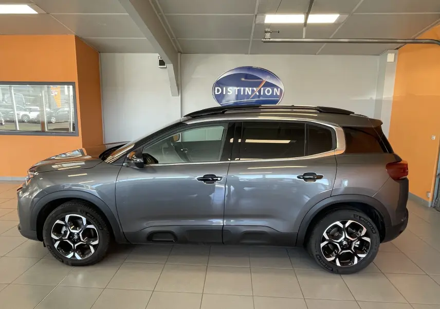 Vue latérale droite d'un Citroën C5 Aircross hybride gris platinium avec jantes alliage biton et barres de toit en intérieur showroom.