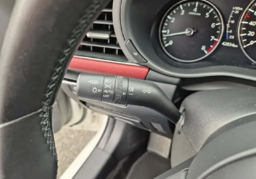 Gros plan sur la manette des feux et clignotants à gauche du volant d'une Mazda3 blanche, avec tableau de bord visible.