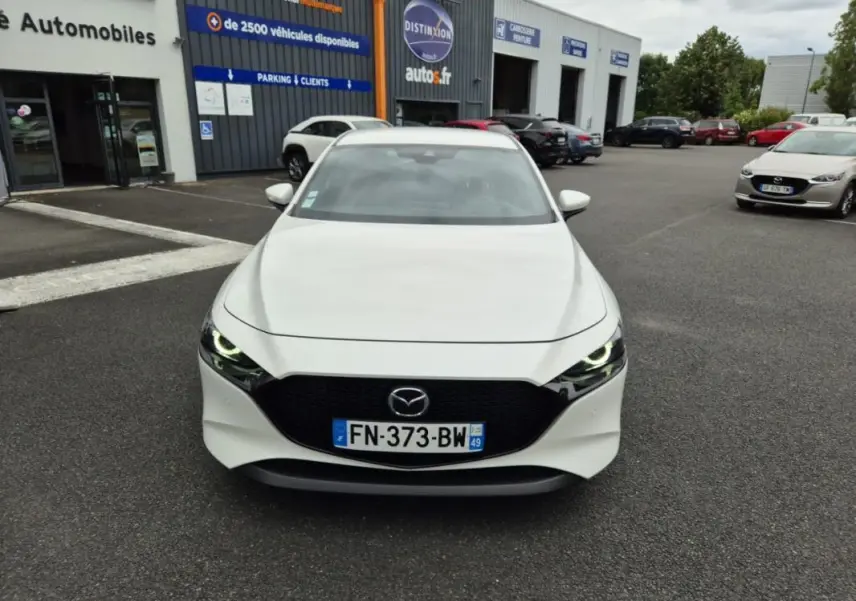 Vue avant d'une Mazda3 blanche de 2020 avec calandre noire et phares allumés sur un parking extérieur.