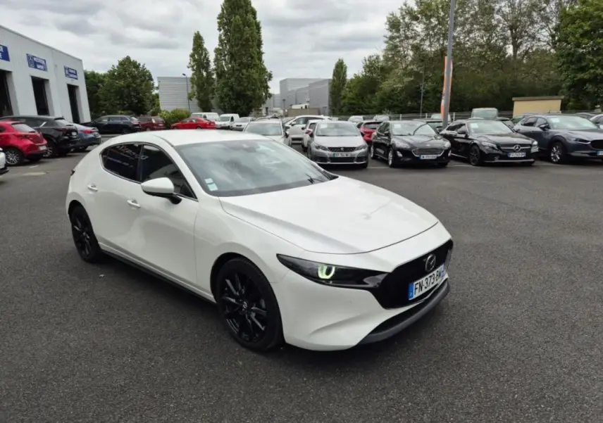 Vue 3/4 avant gauche d'une Mazda3 blanche de 2020 avec jantes noires et phares allumés sur parking.