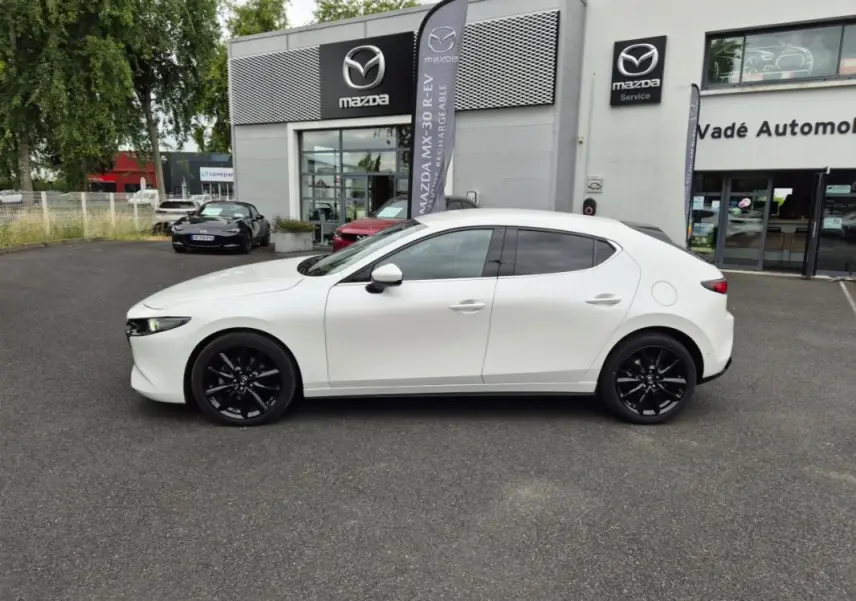 Vue de profil côté gauche d'une Mazda3 blanche avec jantes noires, stationnée devant un concessionnaire Mazda.