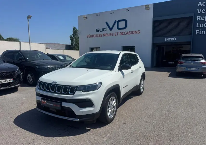 Jeep Compass blanc vue 3/4 avant droit, stationné devant un bâtiment de vente de véhicules.