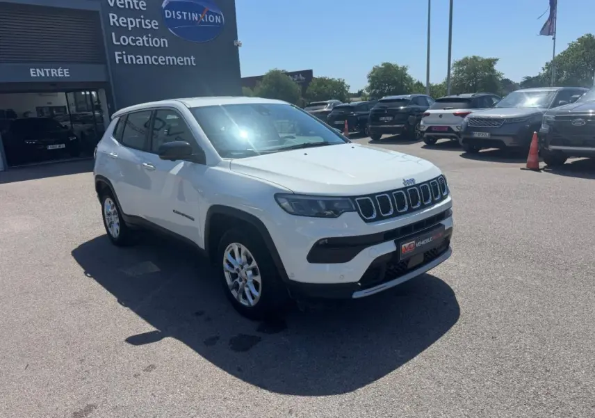Jeep Compass blanc en 3/4 avant droit, stationné devant un bâtiment commercial sous un ciel dégagé.