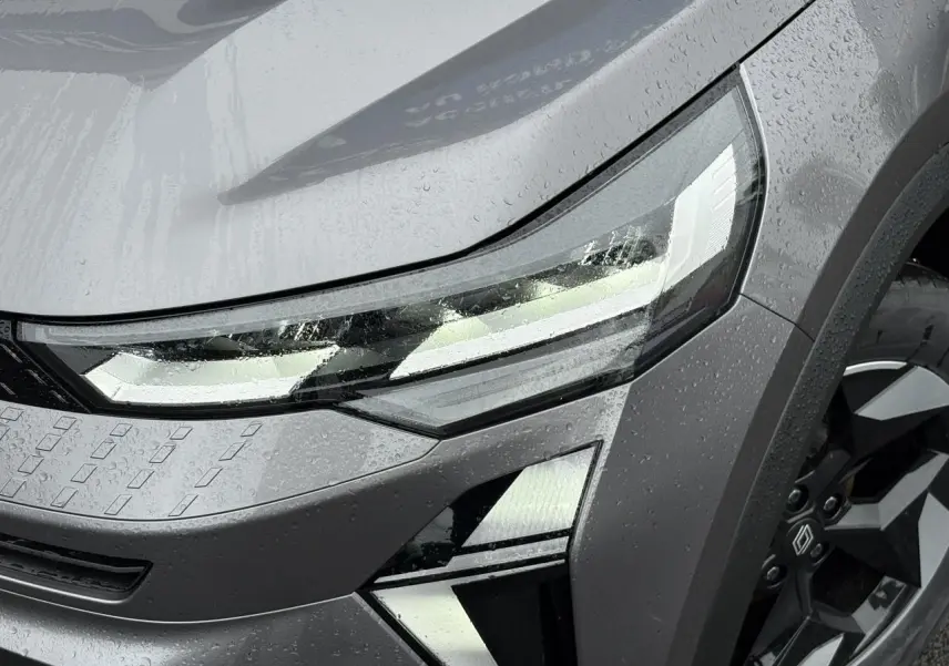 Gros plan sur le phare avant droit et la jante noire du Renault Captur Gris Cassiopée 2025 sous la pluie.
