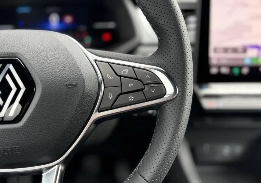 Gros plan sur le volant perforé du Renault Captur 2025 avec commandes intégrées et écran tactile flou en arrière-plan.