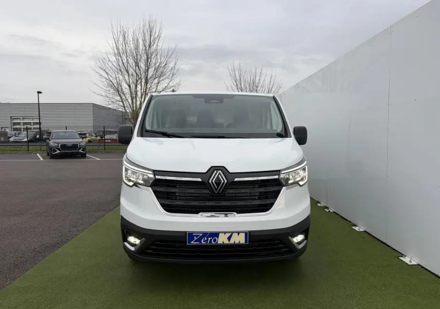 Vue frontale d'un Renault Trafic Fourgon blanc Glacier avec feux LED allumés sur un sol vert synthétique.