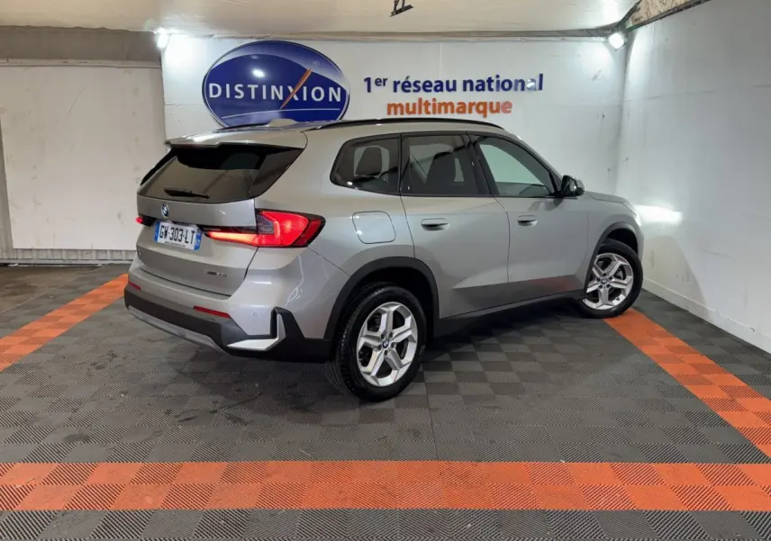 BMW X1 sDrive20i gris foncé vue 3/4 arrière droit dans un showroom avec éclairage mural et logo Distinxion.