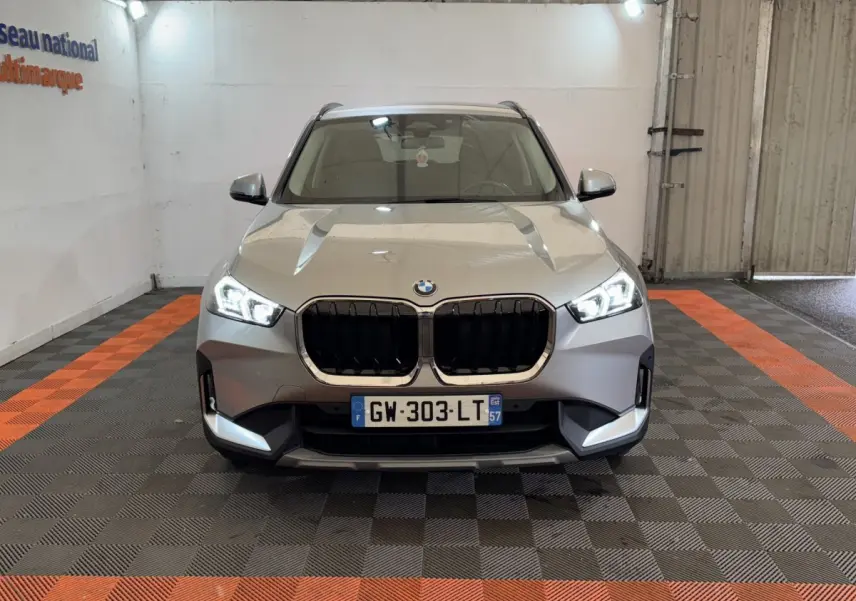 Vue frontale d’un BMW X1 gris foncé 2024 avec calandre noire et phares LED allumés dans un garage.