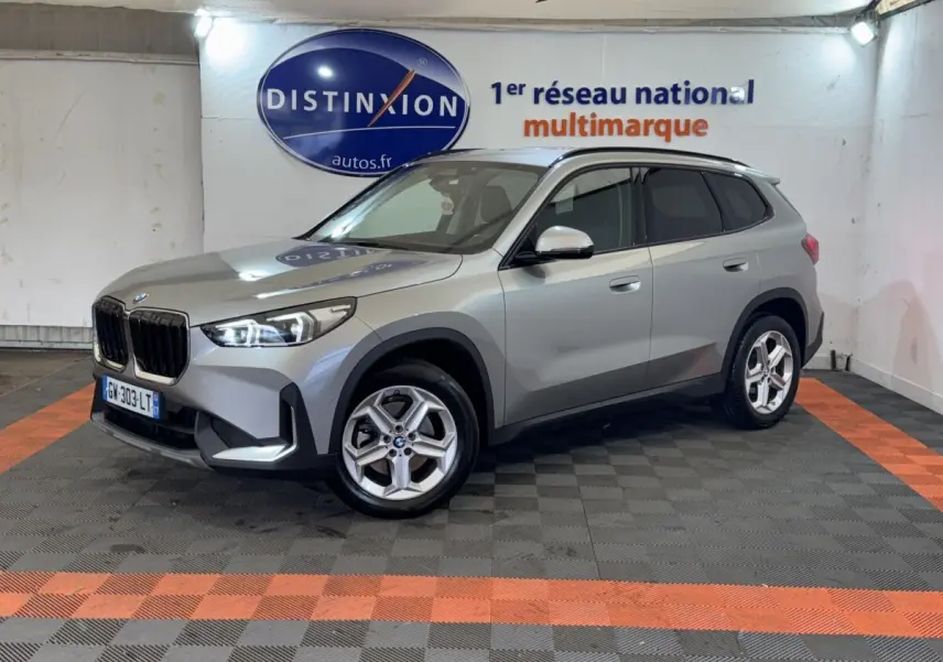 BMW X1 sDrive20i Business Design gris foncé vue 3/4 avant droit dans un showroom avec logo Distinxion en arrière-plan.