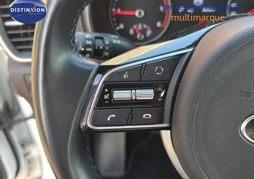 Gros plan sur les commandes gauche du volant noir du Kia Sportage blanc nacré, avec tableau de bord flou en arrière-plan.