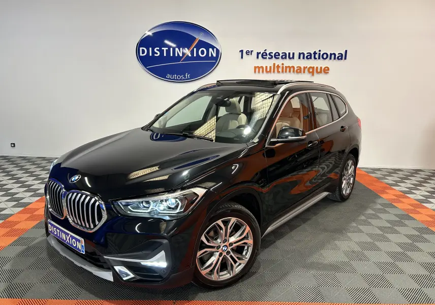 BMW X1 noir en 3/4 avant droit avec toit ouvrant et barres de toit neuves visibles en intérieur showroom.