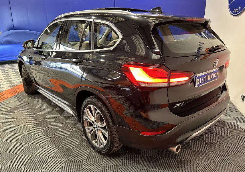 Vue 3/4 arrière droite d'un BMW X1 noir avec feux allumés et barres de toit visibles en intérieur showroom.
