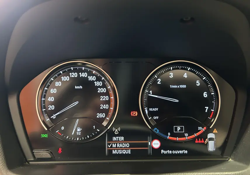 Gros plan sur le tableau de bord numérique du BMW X1 noir, affichant la vitesse, le régime moteur et un message "Porte ouverte".