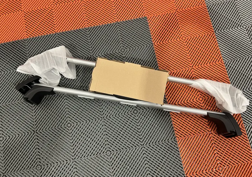 Barres de toit neuves BMW posées au sol sur un sol en damier orange et gris, emballage partiel visible.